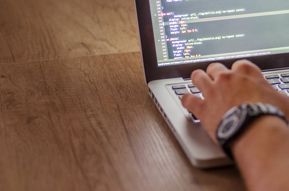 A close-up view of a laptop on a wooden desk displaying lines of code on the screen, with a ceramic mug featuring printed text to the right side. The desk surface appears clean and organized, with sof