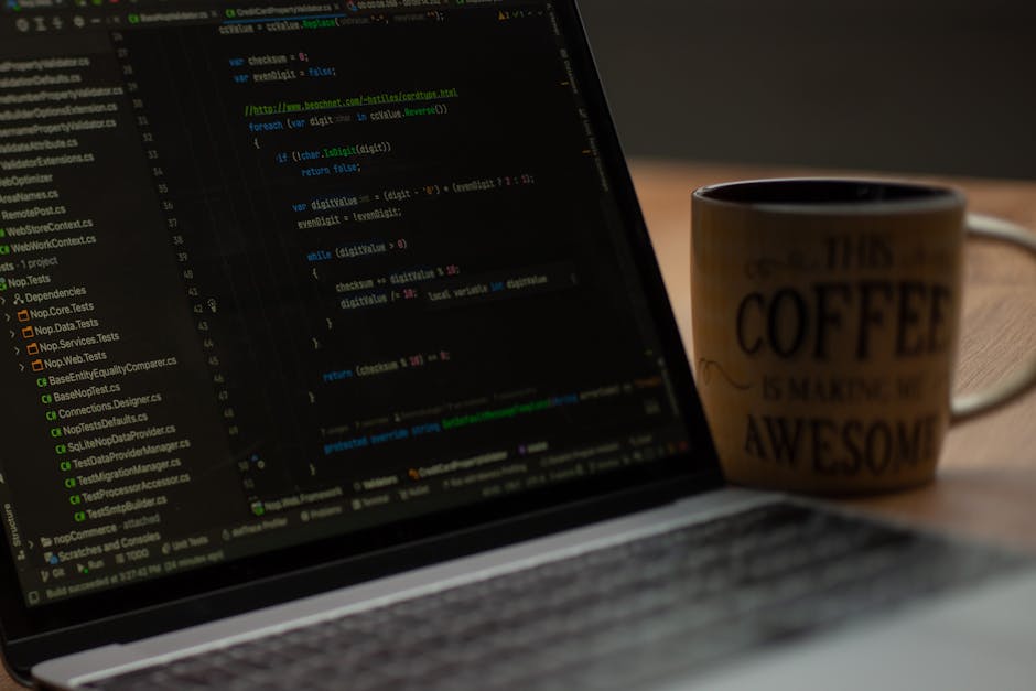 A black and white photograph of a workspace featuring a laptop displaying code on the screen, placed on a dark wooden desk against a textured brick wall. Next to the laptop is an open notebook with a
